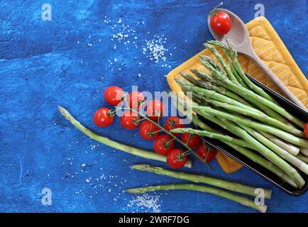 Frischer grüner Spargel und Kirschtomaten auf blau strukturiertem Hintergrund mit Kopierraum. Gesunde Zubereitung des Abendessens. Ausgewogenes Ernährungskonzept. Stockfoto