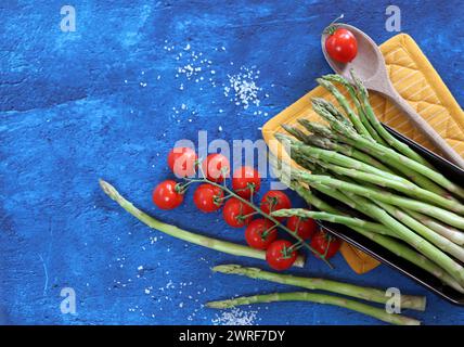 Frischer grüner Spargel mit Kirschtomaten und Meersalz auf blauem Hintergrund Stockfoto