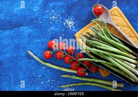 Frischer grüner Spargel und Kirschtomaten auf blau strukturiertem Hintergrund mit Kopierraum. Gesunde Zubereitung des Abendessens. Ausgewogenes Ernährungskonzept. Stockfoto