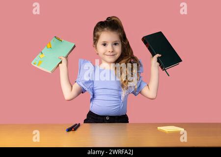 Ein kleines Mädchen, das in einem Schulpult gefangen ist und zwei Bücher hält, die interessante Emotionen zeigen. Das Mädchen liebt es, Bücher zu lesen und zur Schule zu gehen. Hochwertiges Foto Stockfoto