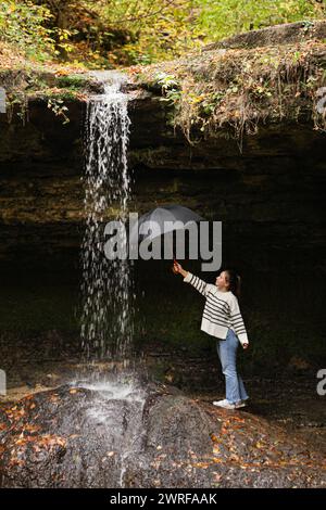 Ein junges Mädchen, das am Morgen aus einer Höhle in einem niedrigen Winkel am Wasserfall steht und einen schwarzen Schirm unter dem fließenden Wasser der Großen hält Stockfoto