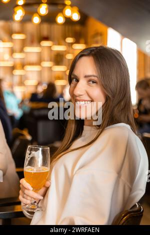 Eine junge, fröhliche Brünette, die dich ansieht, während sie mit einem Glas Bier vor der Kamera gegen Männer, die an der Bar im Pub sitzen, röstet. Stockfoto