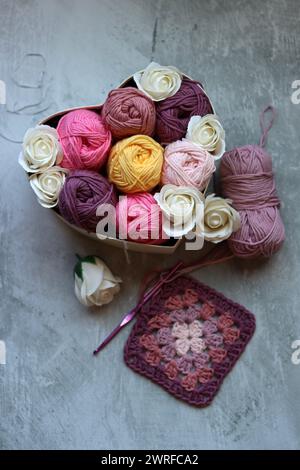 Bunte Kugeln aus Baumwollgarn und weißen Rosen in einem Korb auf grauem Hintergrund mit Platz für Text. Handgefertigte Geschenkbox mit Bio-Garn. Stockfoto