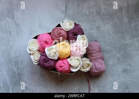 Bunte Kugeln aus Baumwollgarn und weißen Rosen in einem Korb auf grauem Hintergrund mit Platz für Text. Handgefertigte Geschenkbox mit Bio-Garn. Stockfoto