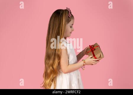 Das Bild des kleinen Mädchens, das in einem weißen Kleid gekleidet ist und ein Geschenk in der Hand hält und sehr begeistert darüber ist, was darin steckt. Bild auf Rosa Stockfoto