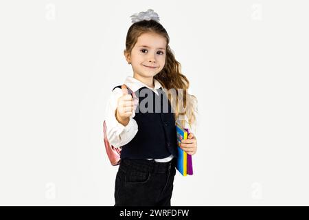 Ein junges Mädchen in formeller Kleidung, mit Blazer und Rucksack, hält ein Buch und gibt mit einem fröhlichen Lächeln die Daumen hoch Stockfoto