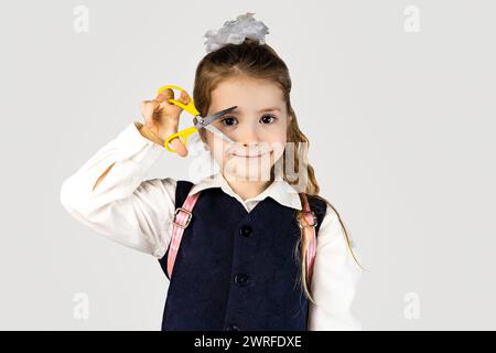 Ein kleines Mädchen in Schuluniform lächelt glücklich und hält eine Schere als Geste für ein Modedesign. Ihre formelle Kleidung besteht aus Kragen und Krawatte Stockfoto