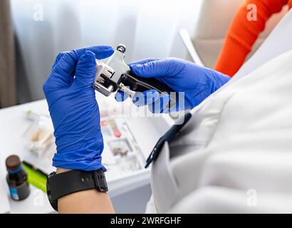 Kosmetikerin lädt die Piercingpistole mit medizinischen Ohrringen in einem Kosmetiksalon auf einem Hintergrund mit Werkzeugen für einen erfolgreichen Piercingprozess. Stockfoto