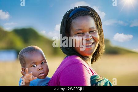 afrikanische Dorfmutter mit Zöpfen trägt das Kind in eine Decke gewickelt im Rücken Stockfoto