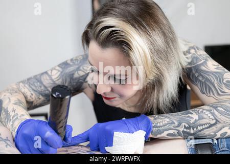 Horizontales Foto der detaillierte Fokus einer Tätowiererin ist spürbar, während sie akribisch an einem Tattoo eines Kunden arbeitet, wobei ihre eigenen farbigen Arme eine Geschichte erzählen Stockfoto