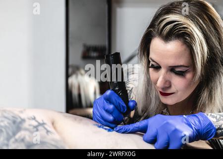 Horizontales Foto eine Nahaufnahme einer Tätowiererin, deren Blick auf die Haut fixiert ist, während ihre Hand die Tätowiermaschine mit fachkundiger Präzision über den Kunden führt Stockfoto