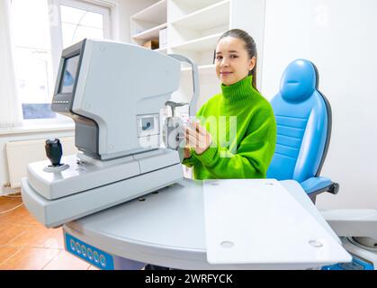 Eine Frau in einem gemütlichen grünen Pullover sitzt bequem an einem Tisch, lächelt vor einem Bürogerät, einer Maschine, die Augenmaße misst, Ophthalmo Stockfoto