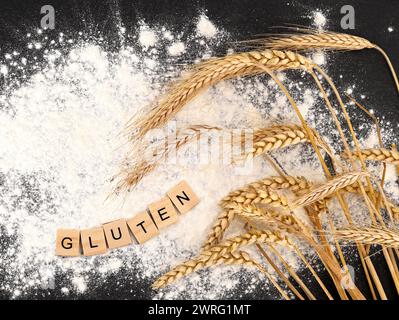 Das Wort Gluten steht in Holzbuchstaben auf Mehl neben Weizenohren auf schwarzem Schiefergrund. Stockfoto