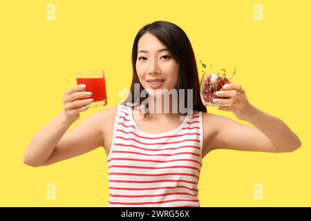 Wunderschöne asiatische Frau mit Kirschen und einem Glas Saft auf gelbem Hintergrund Stockfoto