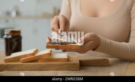 Zugeschnittene Ansicht weibliche Hände verteilen Schokoladenpaste auf Scheiben Toast mit Messer Erdnussbutter auf Brot in Nahaufnahme geben. Unerkennbare Frau in der Küche Stockfoto