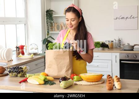 Schöne junge asiatische Frau mit Papiertüte voller frischer Produkte am Tisch in der Küche Stockfoto