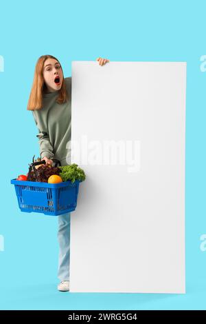 Schockierte junge Frau mit Warenkorb und leerem Poster auf blauem Hintergrund. Preissteigerungskonzept Stockfoto