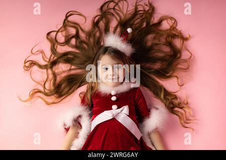 Foto von oben eines traumhaften kleinen Mädchens, das ein weihnachtliches Kleid mit langen blonden Haaren trägt und auf dem Boden liegt und den Weihnachtsgeist genießt. Christma Stockfoto