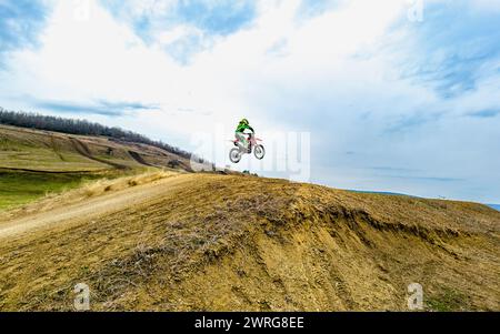 Eine Person mit Helm fährt mit einem Motocross-Bike auf einer unbefestigten Straße und wirft Staub auf, während sie auf dem grasbewachsenen Hang unter bewölktem Himmel rasen Stockfoto