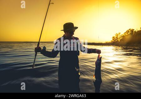 Ein Fischer mit einer Angelrute in der Hand und einem gefangenen Fisch steht im Wasser vor einem wunderschönen Sonnenuntergang. Stockfoto