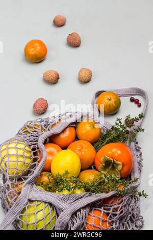 Persimmon, Äpfel und Zitrone im wiederverwendbaren Netzbeutel. Litschi, Preiselbeeren und Mandarinen auf dem Tisch. Grauer Hintergrund. Draufsicht Stockfoto