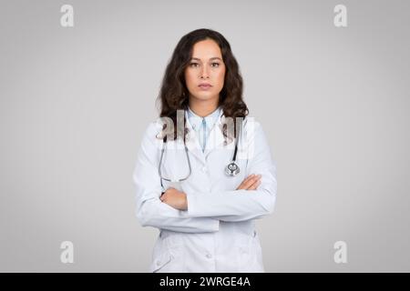 Porträt einer selbstbewussten Ärztin mit gefalteten Armen, die in die Kamera blickt, vor grauem Hintergrund stehend Stockfoto
