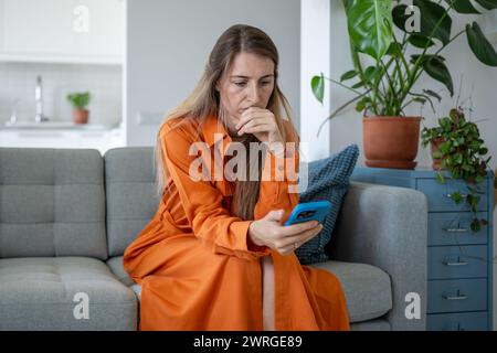 Eine gestresste Frau schaut nervös auf den Smartphone-Bildschirm, drückt die Hand an die Lippen und liest unangenehme Nachrichten. Stockfoto