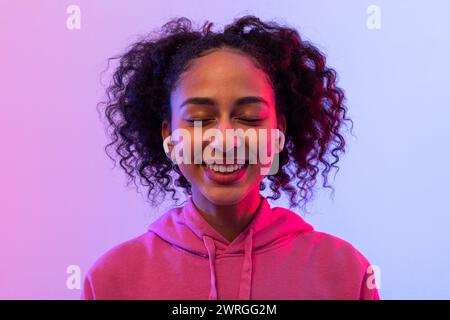 Glückliche schwarze Frau mit lockigem Haar, die Musik in rosa Hoodie und Ohrhörern genießt Stockfoto