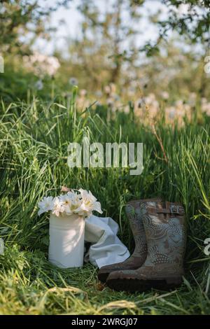 Weiße Narzissen und Tulpen in einer Metalldose und ein Paar Gummistiefel im hohen Gras in einem Frühlingsgarten in Weißrussland Stockfoto