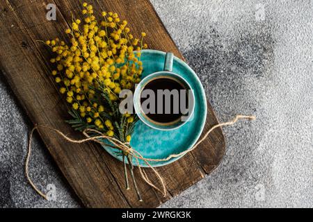 Blick von oben auf eine Tasse schwarzen Kaffee und einen Haufen gelber Mimosa-Blumen auf einem hölzernen Schneidebrett Stockfoto