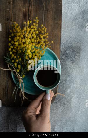 Blick von oben auf eine Frau, die nach einer Tasse schwarzen Kaffee neben einem Haufen gelber Mimosa-Blumen auf einem Tisch greift Stockfoto