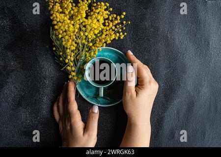 Blick von oben auf eine Frau, die nach einer Tasse schwarzen Kaffee neben einem Haufen gelber Mimosa-Blumen auf einem Tisch greift Stockfoto