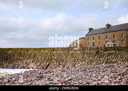 Steinhütte am Hafeneingang Sinclairs Bay, mit dem Meer auf dem Steinschindel und Fischerbooten hinter der Hafenmauer Stockfoto