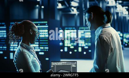 Techniker programmieren im Serverraum mit fortschrittlicher Infrastruktur, die riesige Datensätze speichert. Mitarbeiter, die Code auf einem Laptop ausführen, um Racks in Rechenzentren zu aktualisieren, die komplexe Rechenvorgänge durchführen Stockfoto