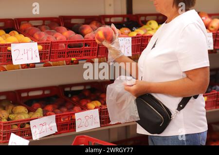 Nicht wiedererkennbare Erwachsene Frau in Kunststoffhandschuhen wählt frischen Pfirsich, kauft Bio-Früchte im europäischen Lebensmittelgeschäft oder Supermarkt. Preise in Euro. Konzept Stockfoto