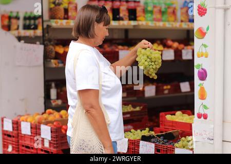 Frau mittleren Alters hält einen Zweig frischer gelber Trauben, wählt und kauft Früchte in einem Supermarkt oder Supermarkt. Das Konzept des Einkaufs-Bio-Lebensmittels Stockfoto