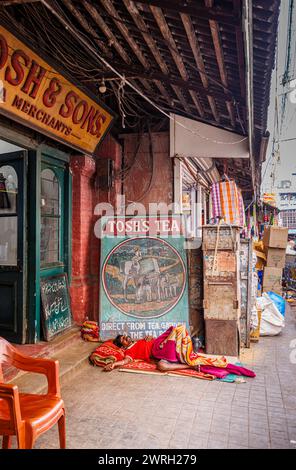 Straßenszene vor dem New Market in Kalkutta, der Hauptstadt von Westbengalen, Indien: Ein einheimischer Mann schläft auf dem Bürgersteig mit einem alten, bemalten Teeschild Stockfoto