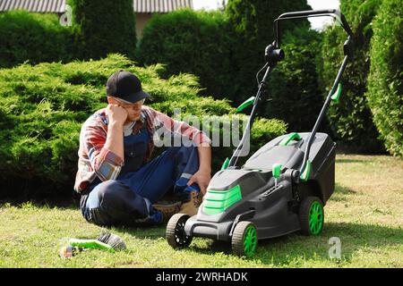 Müder junger Mann, der in der Nähe eines kaputten Rasenmähers im Garten sitzt Stockfoto