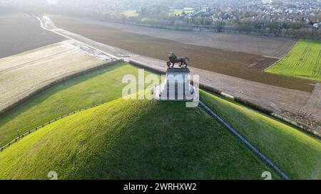 Waterloo, Brüssel, Belgien, 25. Februar 2024 dieses Luftbild zeigt den Löwenhügel am Waterloo Battlefield Memorial. Die Sonne am Horizont strahlt ein warmes Licht über die Landschaft und hebt die Texturen des Grases und der gepflügten Felder hervor. Die Löwenstatue steht auf dem Sockel mit Blick auf die historische Stätte. Der Kontrast zwischen dem üppigen grünen Gras des Hügels und den Brauntönen der umliegenden Felder ist beeindruckend. Die Pfade, die zur Statue und um den Hügel führen, sind deutlich sichtbar, ebenso wie einige Besucher in der Nähe der Basis, was einen Eindruck von der Mo vermittelt Stockfoto