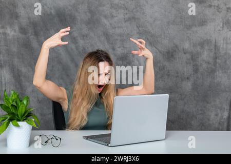Stilvolle junge Frau in grauem Kleid posiert vor Laptop und grüner Blume Stockfoto