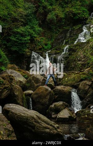 Männlicher Tourist posiert für ein Foto auf Felsbrocken am Torc Waterfall im Killarney National Park, County Kerry, Irland Stockfoto