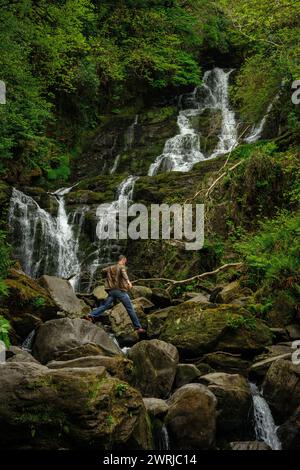 Männliche Touristen springen auf Felsbrocken am Torc Waterfall im Killarney National Park, County Kerry, Irland Stockfoto
