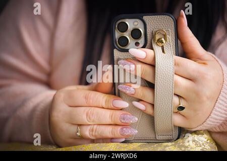 Eine Nahaufnahme der Hände einer Frau, die ein Smartphone hält. Die Frau hat atemberaubende pinkfarbene Gelnägel, sorgfältig bemalt mit zwei Nägeln, die mit Stiften verziert sind Stockfoto