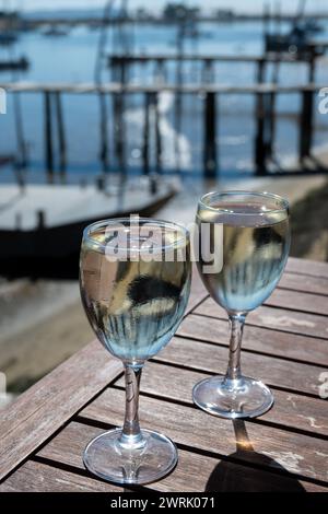 Trinken Sie Weißwein in einem Bauerncafé in einem Dorf mit Austernzucht, mit Blick auf Boote und Wasser der Bucht von Arcachon, Halbinsel Cap Ferret, Bordeaux, Frankreich in Stockfoto