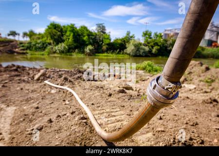 Langer Feuerlöschschlauch mit Metallkupplung an der Flussküste auf der Baustelle zur Wasserversorgung, Brückenbau ist im Gange. Stockfoto