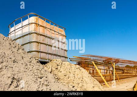 Weißer quadratischer Wassertank, kubisch, mit Metallgitter, riesiger Flüssigkeitsbehälter auf Holzpaletten auf der Baustelle, im Hintergrund ist Brückengerüst-Reinforc Stockfoto