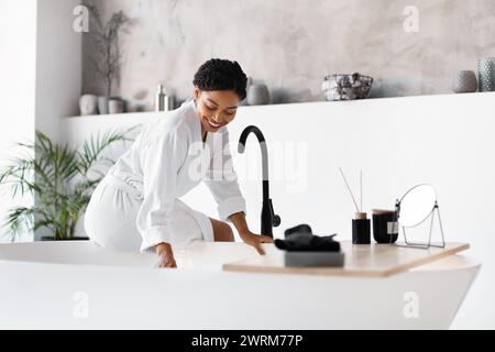 Lächelnde schwarze Frau im Bademantel, die sich über moderne freistehende Badewanne zu Hause lehnt Stockfoto