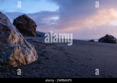 Ein felsiger Strand mit einem großen Felsen im Vordergrund. Der Himmel ist bewölkt und die Sonne untergeht Stockfoto