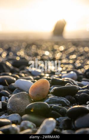 Ein kleiner Felsen sitzt an einem Strand mit einem großen Felsen im Hintergrund. Der Fels ist von vielen anderen Felsen umgeben, was eine felsige und zerklüftete Landschaft schafft Stockfoto