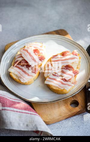 Von oben zwei Toasts bedeckt mit dünn geschnittenen Schmalzstücken, präsentiert auf einer Keramikplatte mit Holzbrett und Stoffserviette. Stockfoto